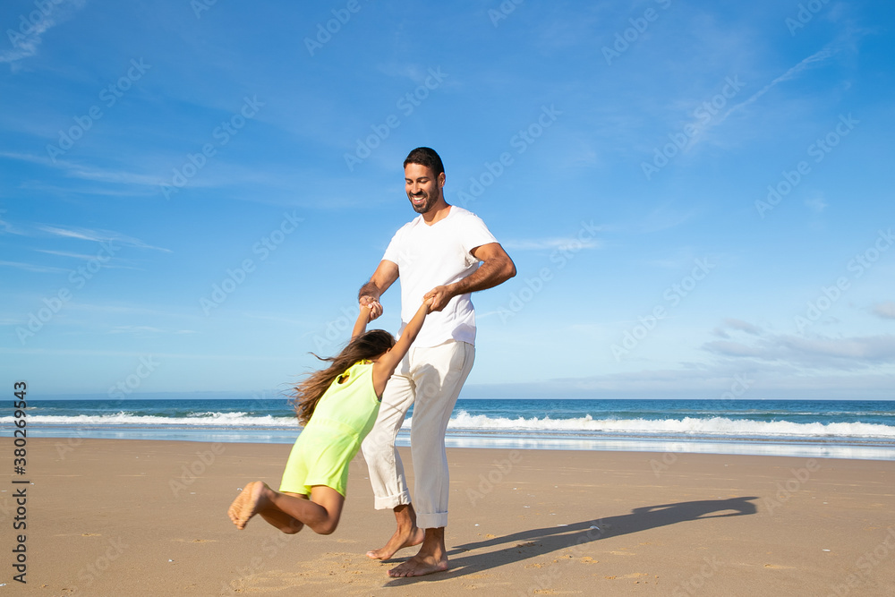 Joyful dad and daughter having fun on beach, playing active games. Father holding girls hands and spinning around. Full length. Family outdoor activities concept