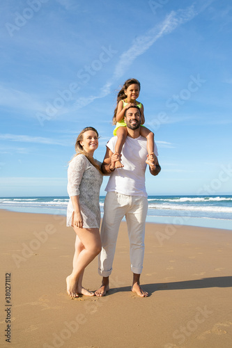 Joyful parents and child wearing summer clothes, posing on ocean beach, girl riding on dads neck. Vertical shot. Family on summer vacation at sea concept