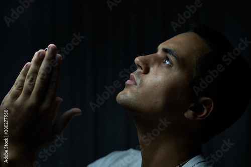 brunette boy in white t-shirt on black background praying. teenager looking at god with serious gaze and low light. man in silver necklace with serious expression while begging