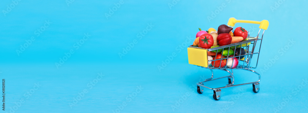 Fototapeta premium Metal shopping cart with fruits and vegetables on a blue background. Toy miniature shopping trolley