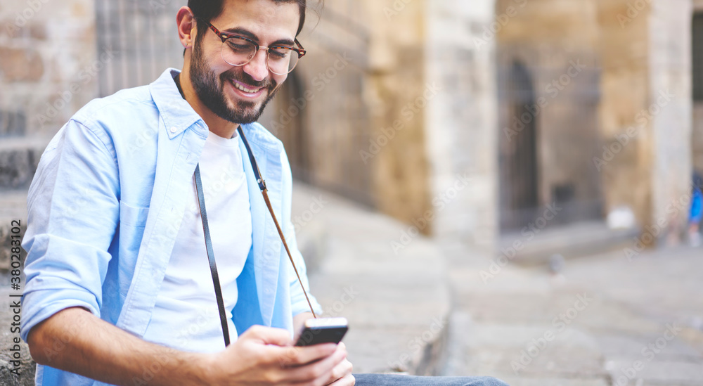 Cheerful young unshaven man in glasses sitting on stairs and browsing smartphone Cheerful young unshaven man in glasses sitting on stairs and browsing smartphone