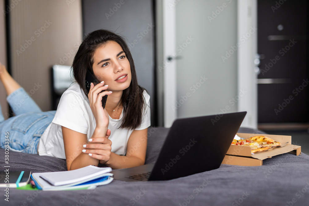 Young woman sitting on the bed with her laptop and talking on the phone with box of pizza Young woman sitting on the bed with her laptop and talking on the phone with box of pizza