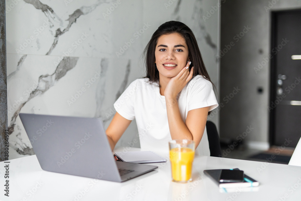 © F8  \ Suport Ukraine - Young woman typing on laptop and smiling while drinking juice in white kitchen