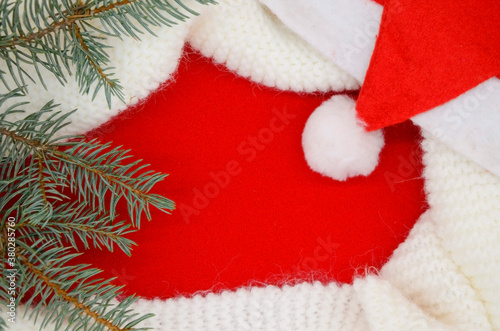 White knitted scarf and Santa hat, Christmas tree branch on red background. © Geparda