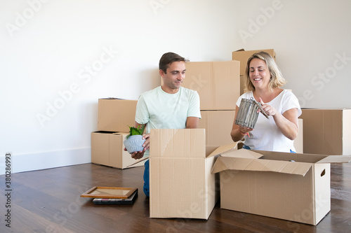 Cheerful married couple moving into new apartment, unpacking things, sitting on floor and taking objects from open boxes. Copy space. Relocation or moving concept