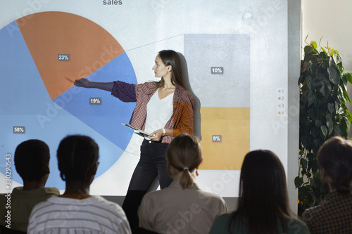 Side view portrait of young woman pointing at data chart with statistics while giving presentation during business conference, copy space photo