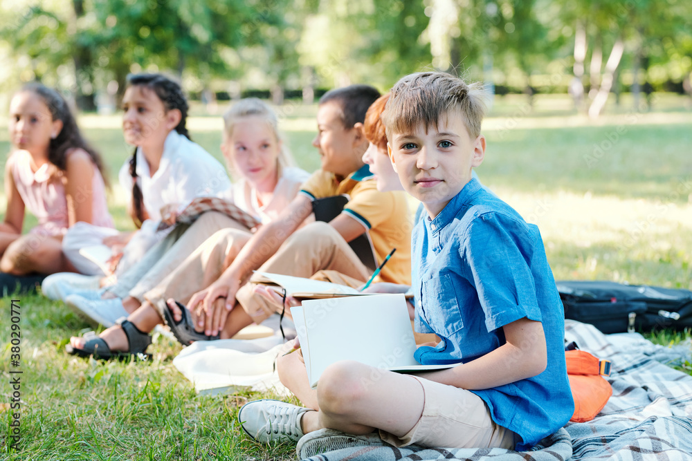 Portrait of content boy in blue shirt sitting with crossed legs on plaid together with classmates and holding workbook at outdoor school lesson