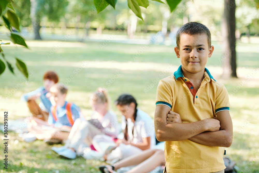 Portrait of smiling confident boy in yellow tshirt standing with crossed arms in summer park, student doing task in background Portrait of smiling confident boy in yellow tshirt standing with crossed arms in summer park, student doing task in background