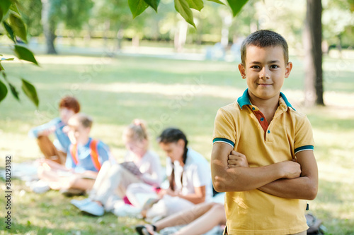 Portrait of smiling confident boy in yellow tshirt standing with crossed arms in summer park, student doing task in background