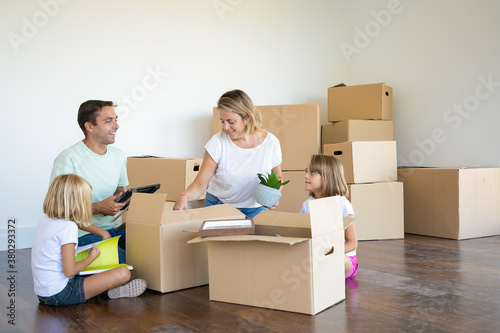 Parents and kids unpacking things in new apartment, sitting on floor and taking objects from box. Relocation or moving concept © Mangostar