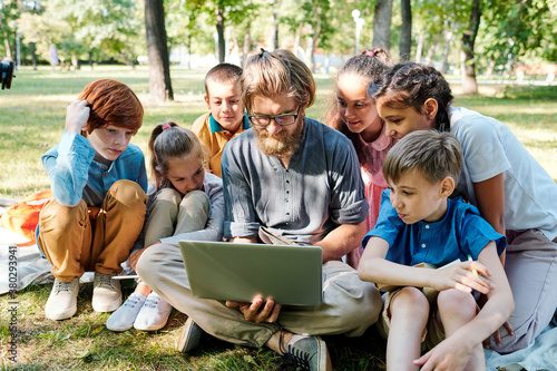 Young bearded teacher in eyeglasses sitting on grass and using laptop while showing pictures to kids at outdoor class