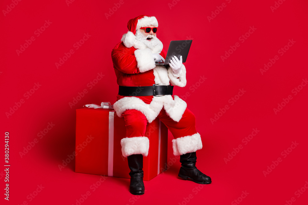 Full length photo of retired old man white beard sit giftbox hold laptop take orders online wear x-mas santa costume glove coat belt sunglass cap boot isolated red color background Full length photo of retired old man white beard sit giftbox hold laptop take orders online wear x-mas santa costume glove coat belt sunglass cap boot isolated red color background