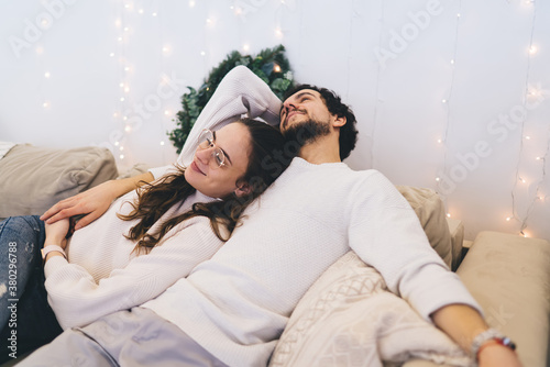 Positive caucasian woman lying near lovely boyfriend satisfied with winter vacations holidays at cozy decorated apartment, romantica couple in love feeling happiness together during christmas evening