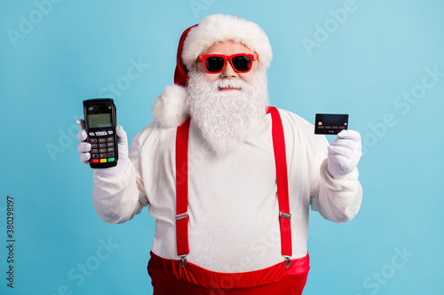 Photo of pensioner old man grey beard hold debit card terminal courier offer cashless pay wear santa x-mas costume suspenders sunglass white gloves cap isolated blue color background