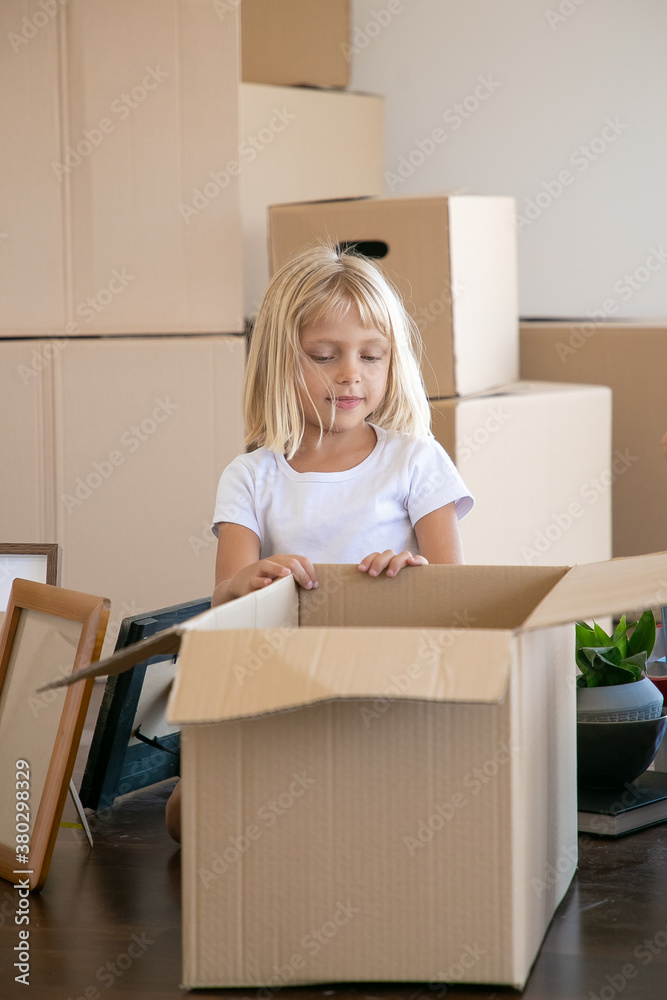 Sweet fair hair little girl unpacking things in new apartment, sitting on floor near open cartoon box and looking inside. Family in new apartment concept