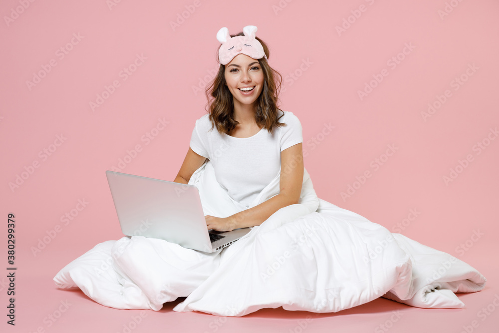 Cheerful young woman in pajamas home wear sleep mask sitting working on laptop pc computer while resting at home isolated on pastel pink background studio portrait. Relax good mood lifestyle concept. Cheerful young woman in pajamas home wear sleep mask sitting working on laptop pc computer while resting at home isolated on pastel pink background studio portrait. Relax good mood lifestyle concept.