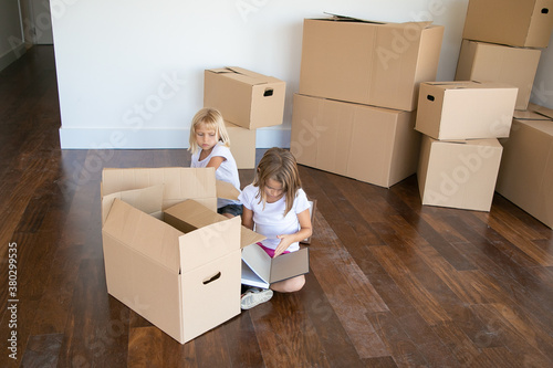Two focused girls unpacking things in new apartment  sitting on floor near open cartoon boxes  holding book. Copy space. Relocation or moving concept