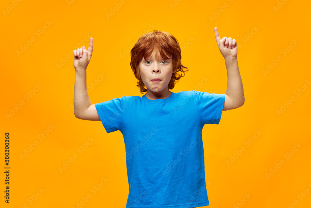 Red-haired boy grimaces face shows two hands up blue t-shirt yellow background 