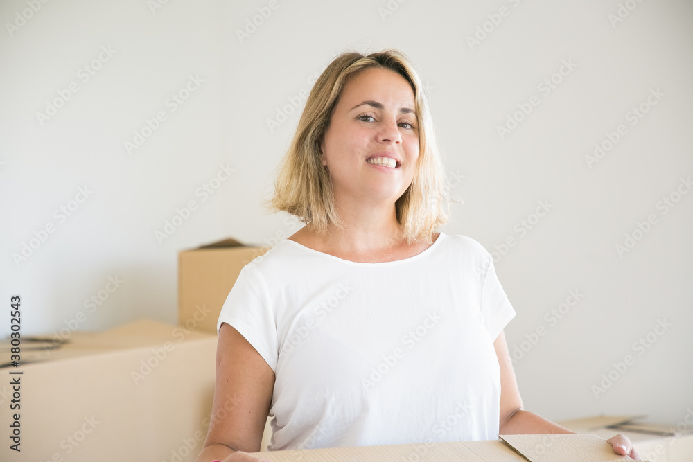 Pretty blonde woman standing with box in new house or apartment. Portrait of attractive Caucasian lady holding cardboard box and smiling. Mortgage, relocation and moving day concept Pretty blonde woman standing with box in new house or apartment. Portrait of attractive Caucasian lady holding cardboard box and smiling. Mortgage, relocation and moving day concept