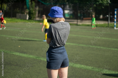 The girl takes pictures of sports. The girl is filming a football match. Girl in shorts on the playground. © Олег Копьёв