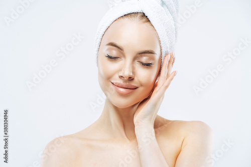 Pretty calm woman with wrap towel on head, closes eyes, enjoys softness of skin, undergoes skin care treatment, stands bare shoulders indoor, relaxed after bath, isolated over white background