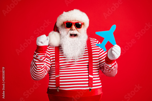 Photo of grandfather grey beard hold paper plane raise fist open mouth wear santa claus x-mas costume suspenders sunglass striped shirt cap isolated red color background © deagreez