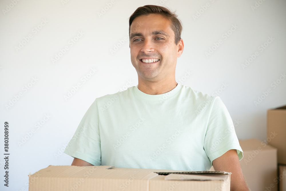Portrait of Caucasian man carrying carton box and smiling. Cheerful handsome home owner standing in new house or apartment and holding cardboard box. Mortgage, relocation and moving day concept Portrait of Caucasian man carrying carton box and smiling. Cheerful handsome home owner standing in new house or apartment and holding cardboard box. Mortgage, relocation and moving day concept