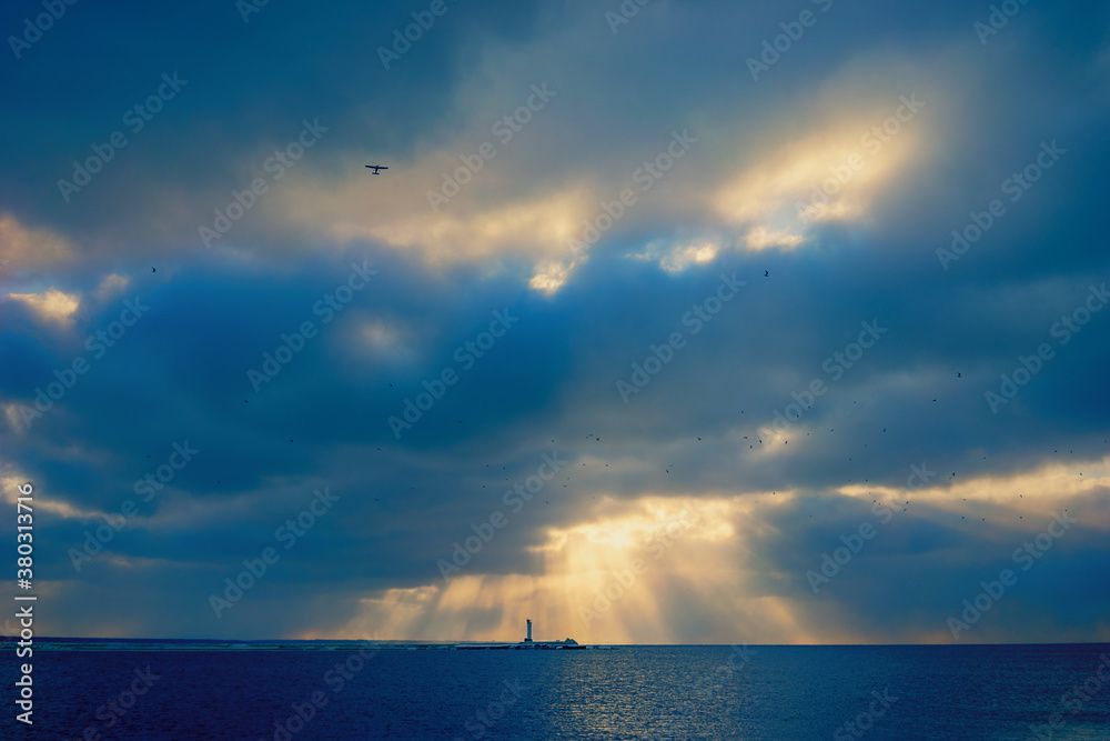 A light plane flies in the blue blue sky among the birds over the sea. Orange sun rays shine through blue clouds against the backdrop of the lighthouse in the bay. Latvia