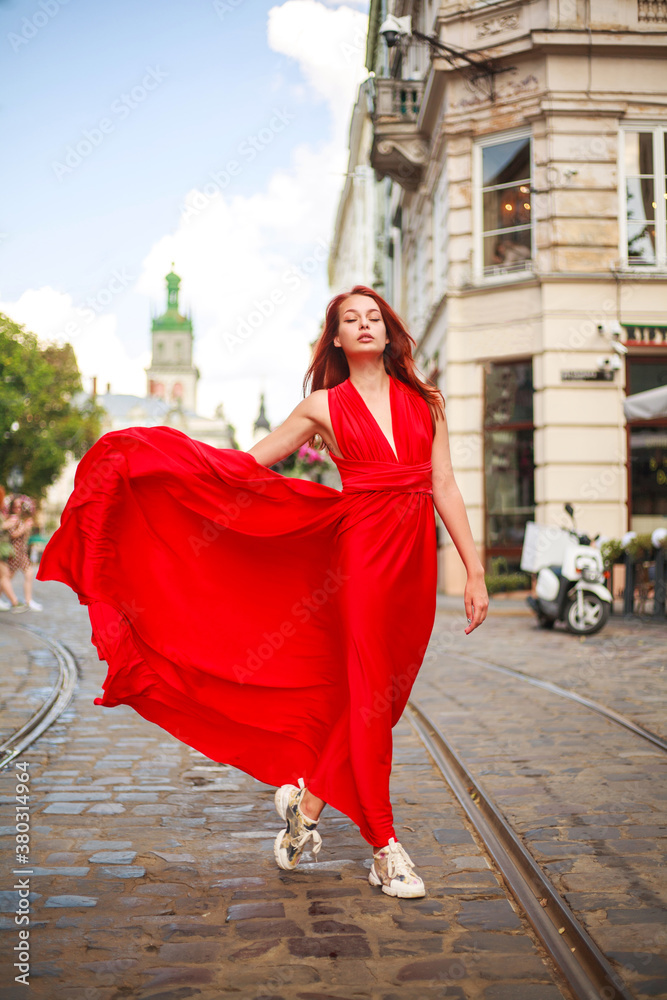 Fototapeta premium Attractive young woman in long red dress is walking along the square of tourist town