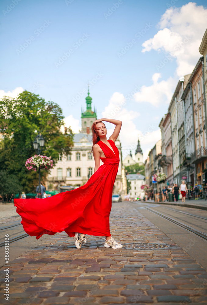 Naklejka premium Attractive young woman in long red dress is walking along the square of tourist town