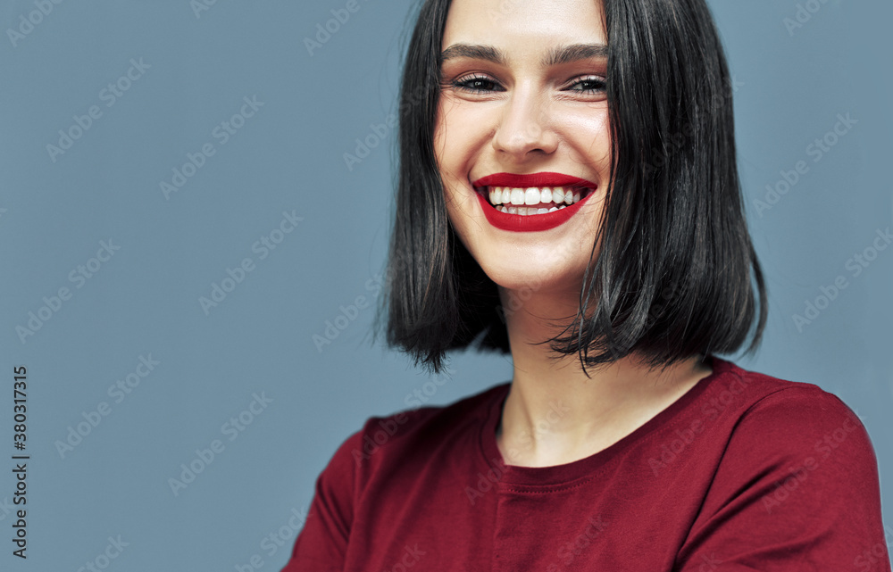 Closeup studio portrait of cheerful young woman with red lips smiling broadly with white teeth. Brunette female model with makeup has joyful expression on blue background. Closeup studio portrait of cheerful young woman with red lips smiling broadly with white teeth. Brunette female model with makeup has joyful expression on blue background.