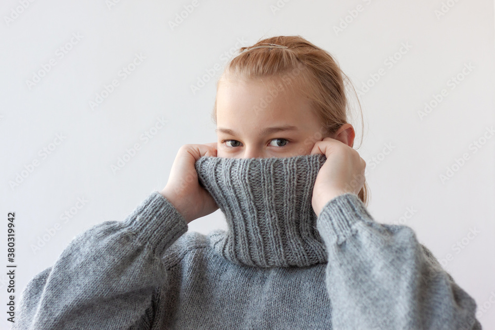 A teenage girl with blonde hair pulled the collar of a gray sweater over her face with her hands, only her eyes are visible A teenage girl with blonde hair pulled the collar of a gray sweater over her face with her hands, only her eyes are visible