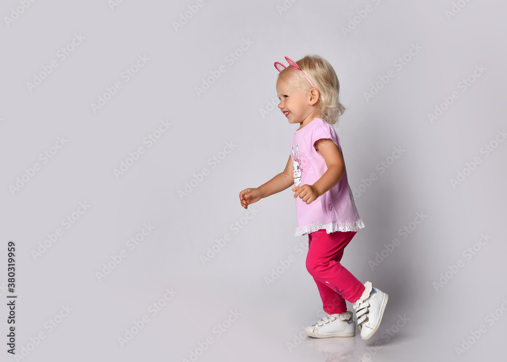 Small cute blonde smiling baby girl in pink stylish casual clothing and kitty ears running towards free copy space Small cute blonde smiling baby girl in pink stylish casual clothing and kitty ears running towards free copy space