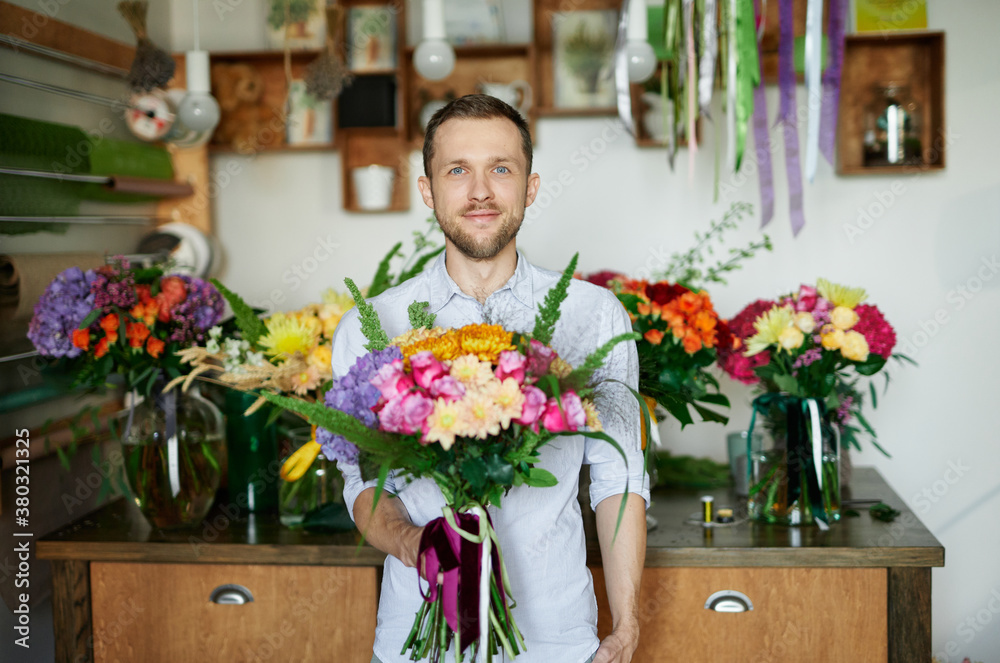 Fototapeta premium Attractive young caucasian man in flower store with bouquet. Good looking male florist in shirt holding bouquet and showing it. Small business concept.