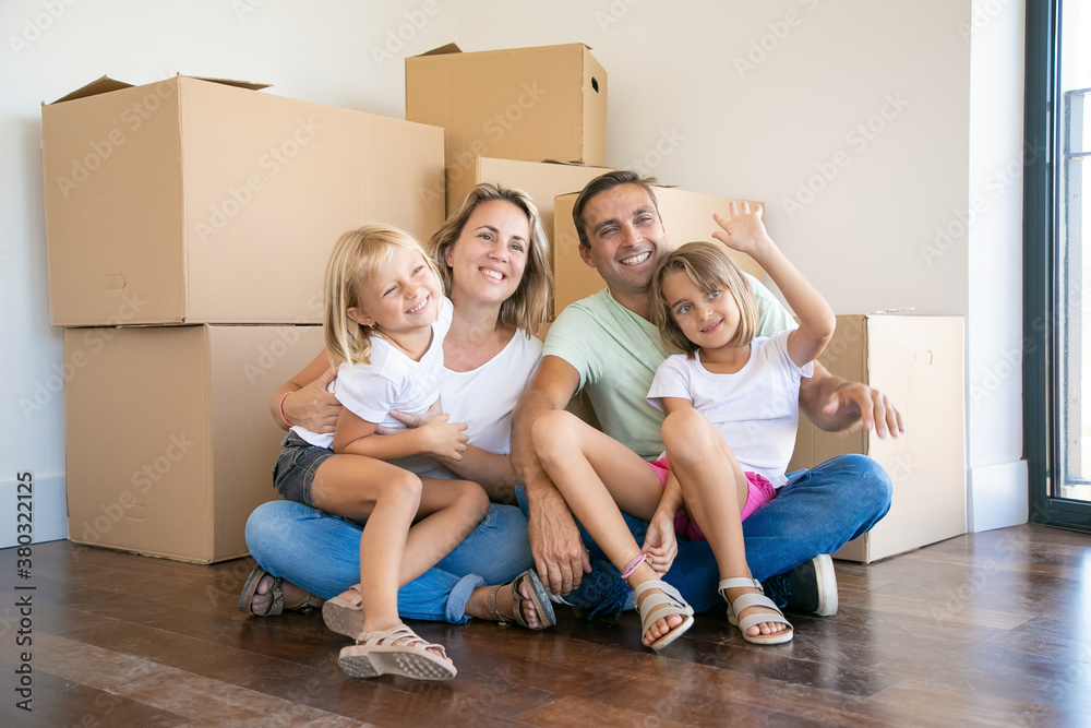 Smiling family with kids sitting on floor near cardboard boxes and relaxing. Blonde girl on father legs waving. New home owners enjoying apartment. Mortgage, relocation and moving day concept Smiling family with kids sitting on floor near cardboard boxes and relaxing. Blonde girl on father legs waving. New home owners enjoying apartment. Mortgage, relocation and moving day concept