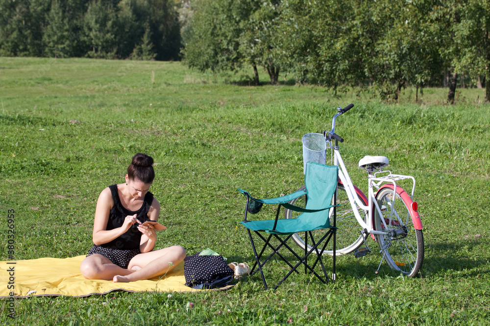 The girl is resting in the park. Sits on the rug, looks at the phone. There is a bicycle and a camping chair nearby.