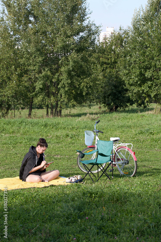 The girl is resting in the park. Sits on the rug, looks at the phone. There is a bicycle and a camping chair nearby.