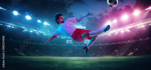 Kicking ball in high jump. Football or soccer player on full stadium and flashlights background. Flyer with copyspace in neon colors. Concept of sport, competition, winning, action and motion.