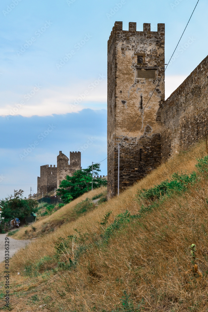 old ancient stone wall and towers of Genoese fortress in Sudak made of sand brown colored bricks stand on steep slope with dry yellow grass. Crimea, summer. Blue sky with clouds. Historic place