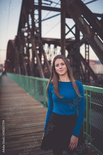 Beautiful young woman with long healthy dark blonde hair in blue long sleeve shirt and black skirt on railway station, looking at the camera. Urban sunset portrait. Late evening sunset time