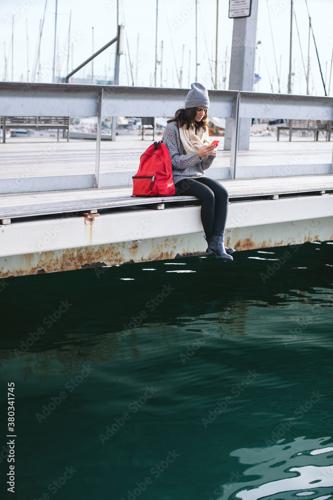 Young beautiful woman using mobile phone wearing winter clothes on seaport.