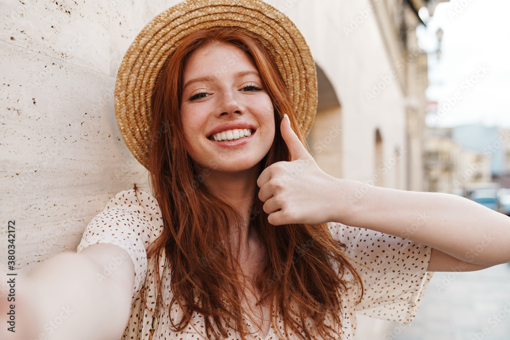 Image of joyful ginger girl showing thumb up while taking selfie