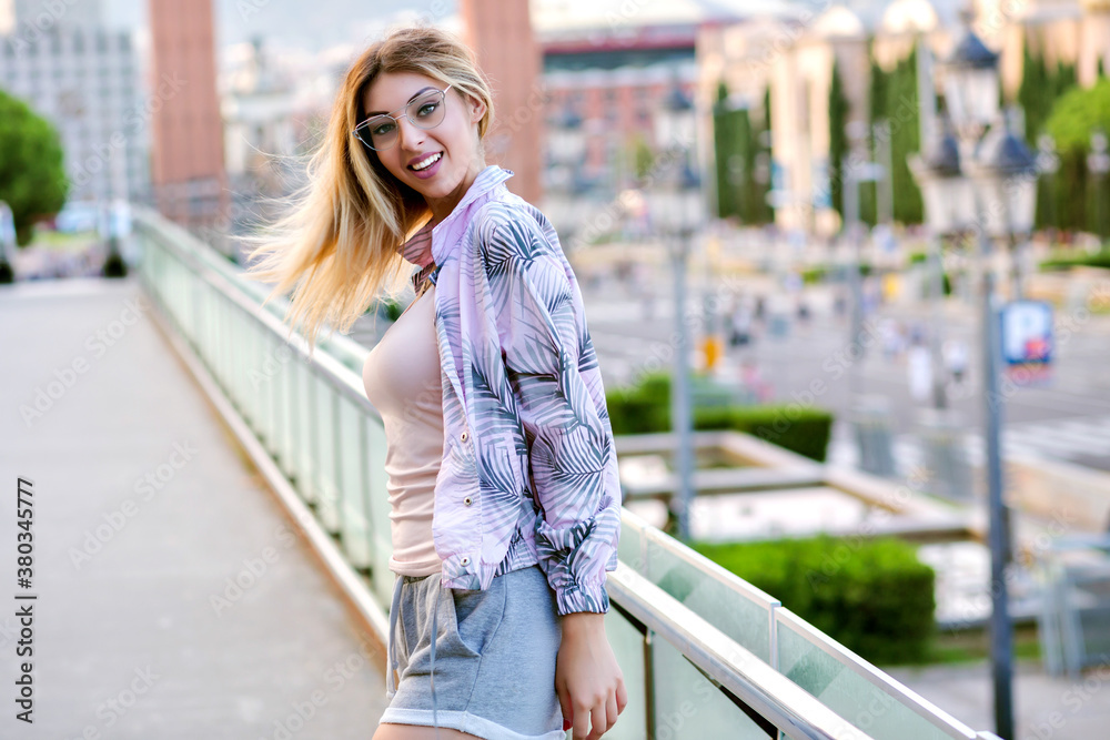 Outdoor positive spring portrait of happy blonde stylish woman, wearing trendy clothes for sport fitness and run, posing at European city square, pastel soft colors.