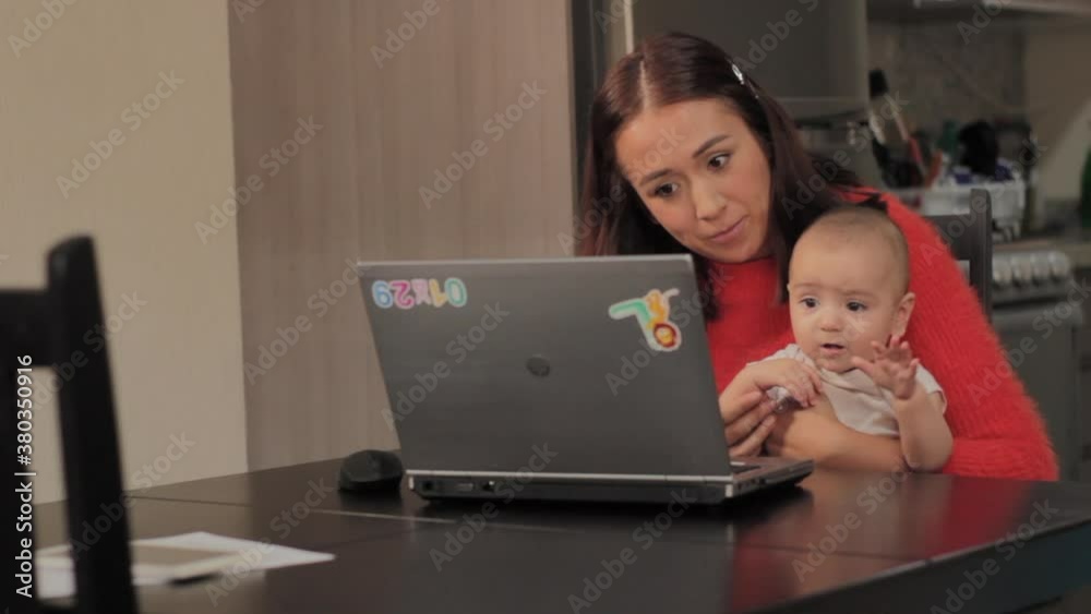 Mujer joven mexicana trabajando desde casa con computadora en comedor dando de comer a bebe
