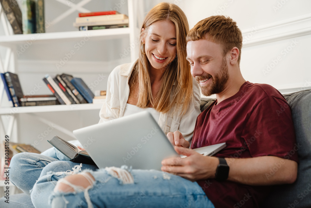 Image of couple reading book and using laptop while sitting on sofa Image of couple reading book and using laptop while sitting on sofa