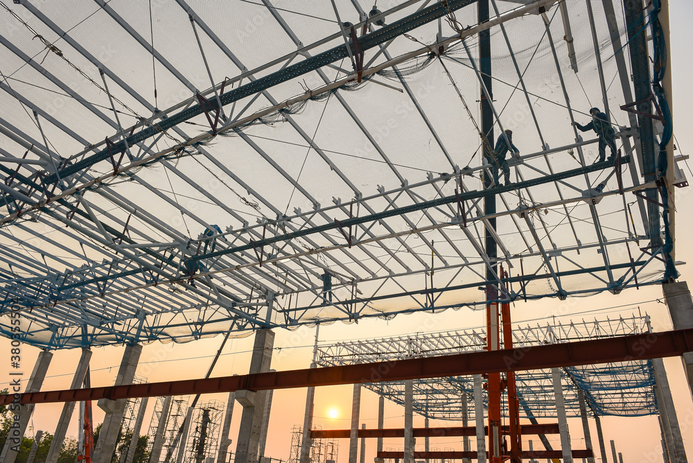 Construction workers are working at height above roof to instrallation accessory steel structure roof truss at construction site factory project during sunset time