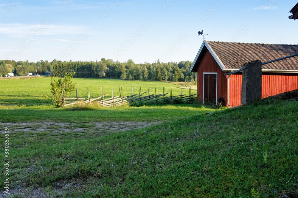 Obraz premium Traditional Finnish red wooden barn with roundpole fence and the weather vane on the roof. Old idyllic farm in the countryside.