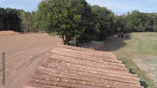 Camera flying above a pile of logs and moving backwards. Green trees and an arid landscape on a summer day are visible. In the background there is a tractor and some hay bales.