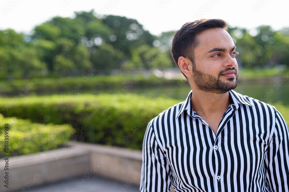Fototapeta premium Young bearded Indian businessman relaxing at the park in the cit