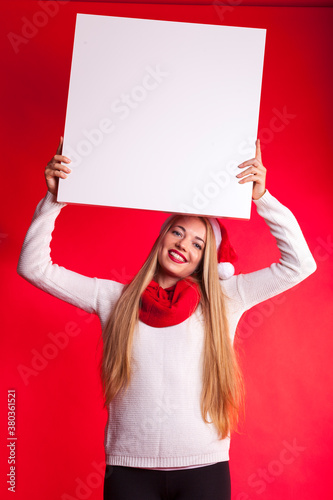 friendly smiling woman with santa hat in christmas mood holding blank white sign board. isolated against red background